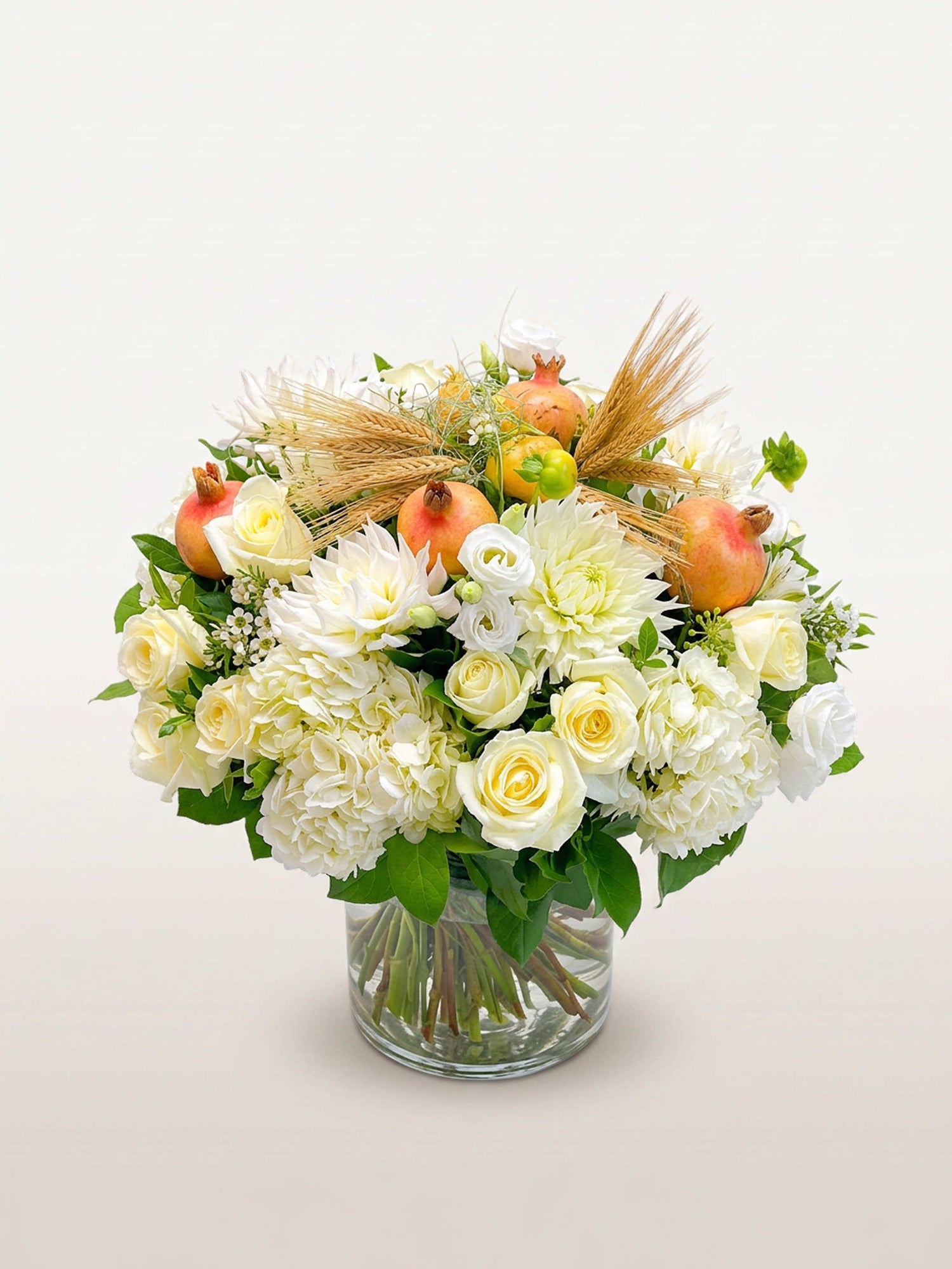 Roses blanches, dahlias, grenades et épis de blé disposés en dôme dans un vase en verre