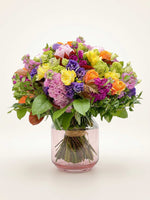 Bouquet rond XL aux teintes vives de rose, violet, jaune et orange, présenté dans un vase en verre rose