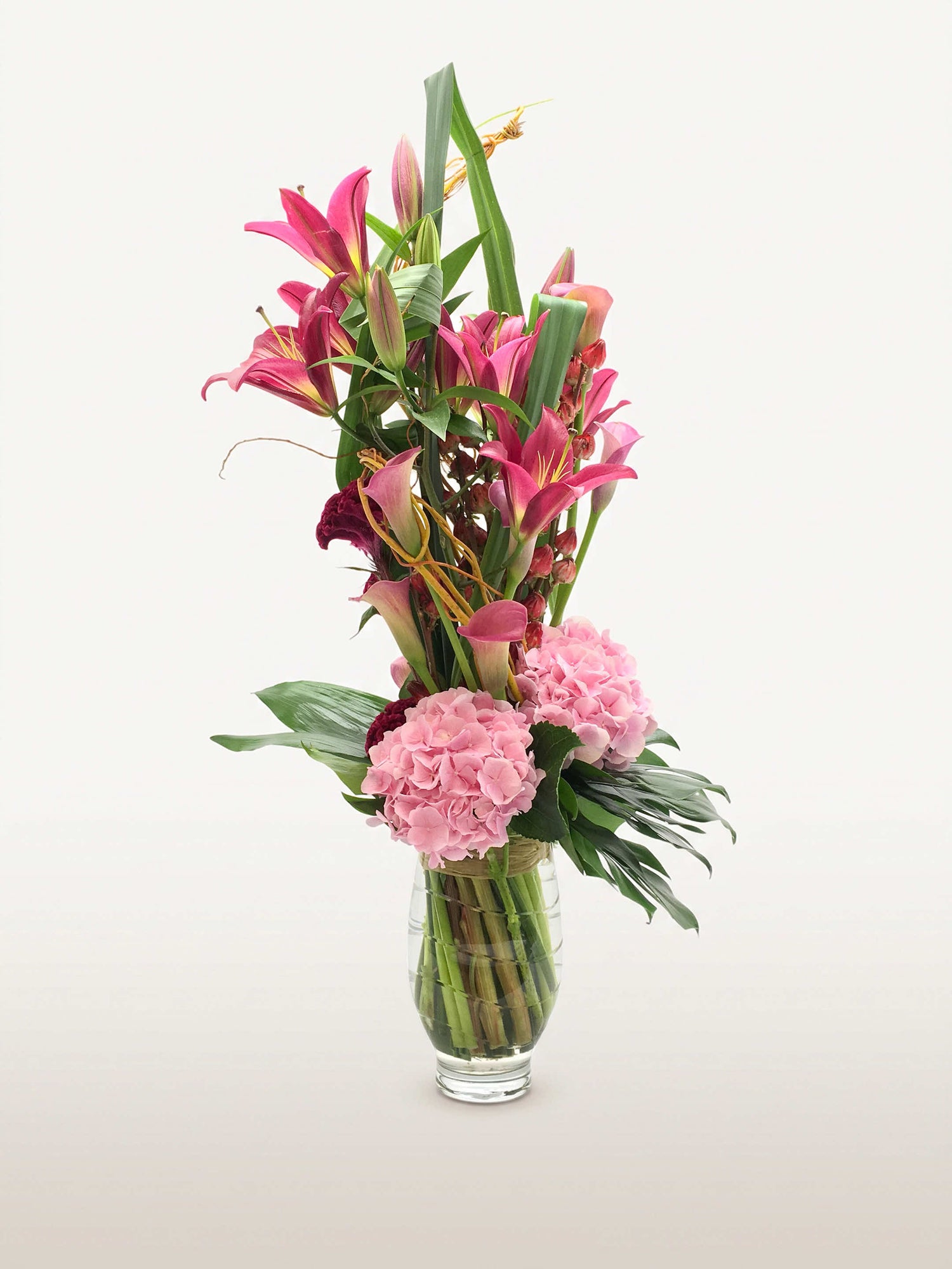 Beaux Arts, lys roses, hortensias roses, callas et feuillages verts dans un vase en verre cylindrique