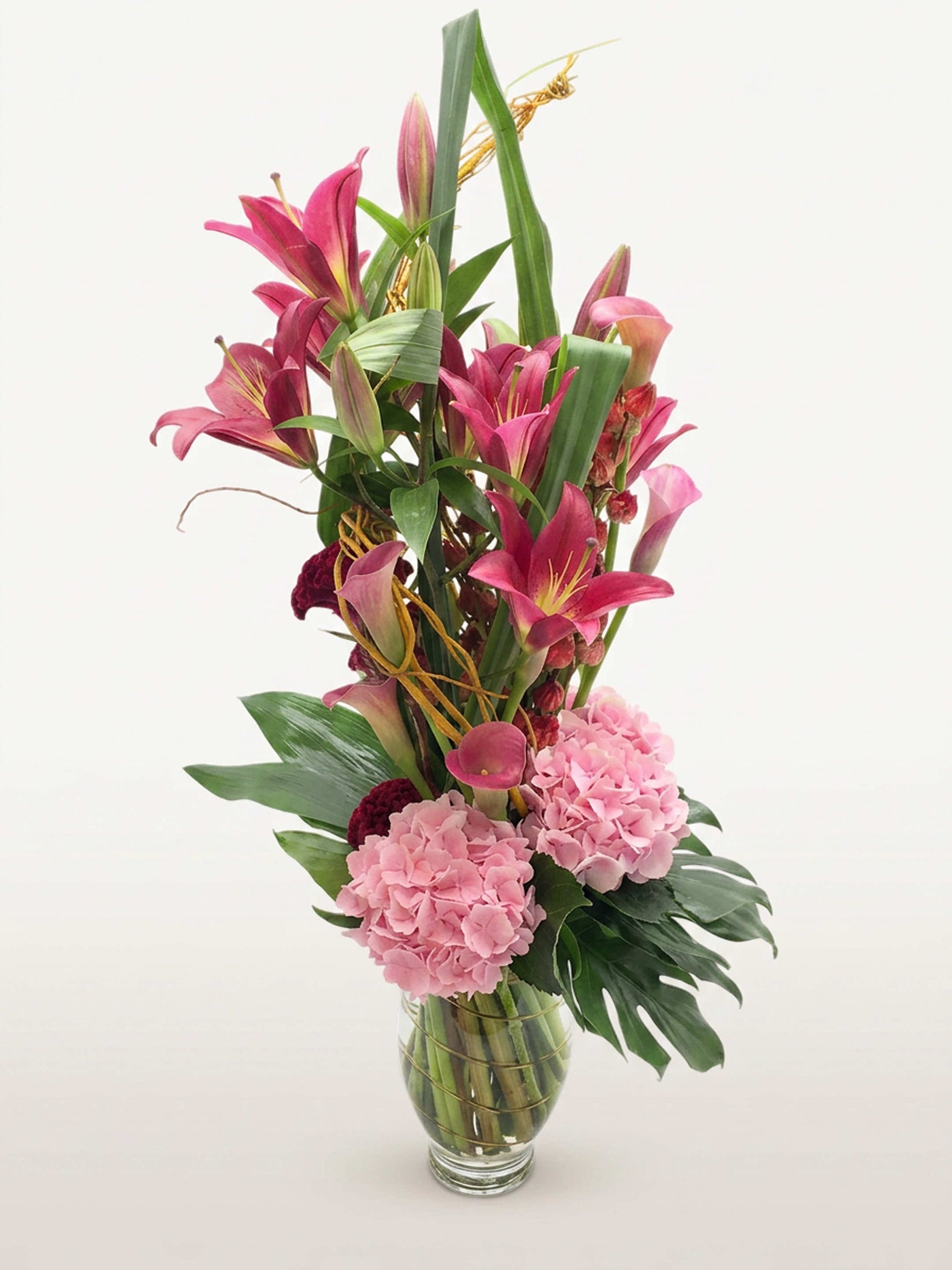 Beaux Arts : lys roses, arums, hortensias roses et feuillages verts dans un vase transparent