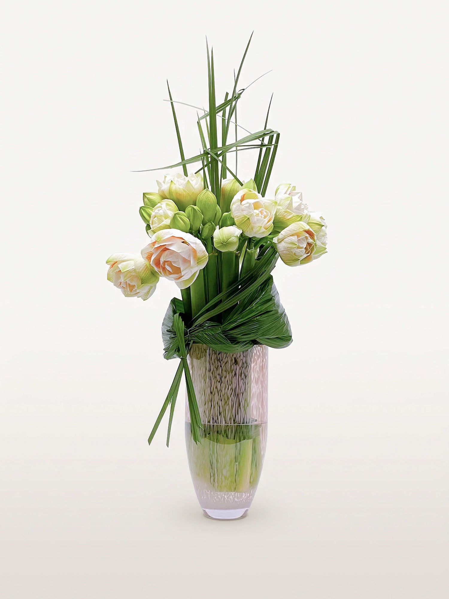 Villa des Nymphéas L déploie d'amarylis blanc-crème et pêche parmi des brins d'herbe graphiques dans un vase en verre translucide