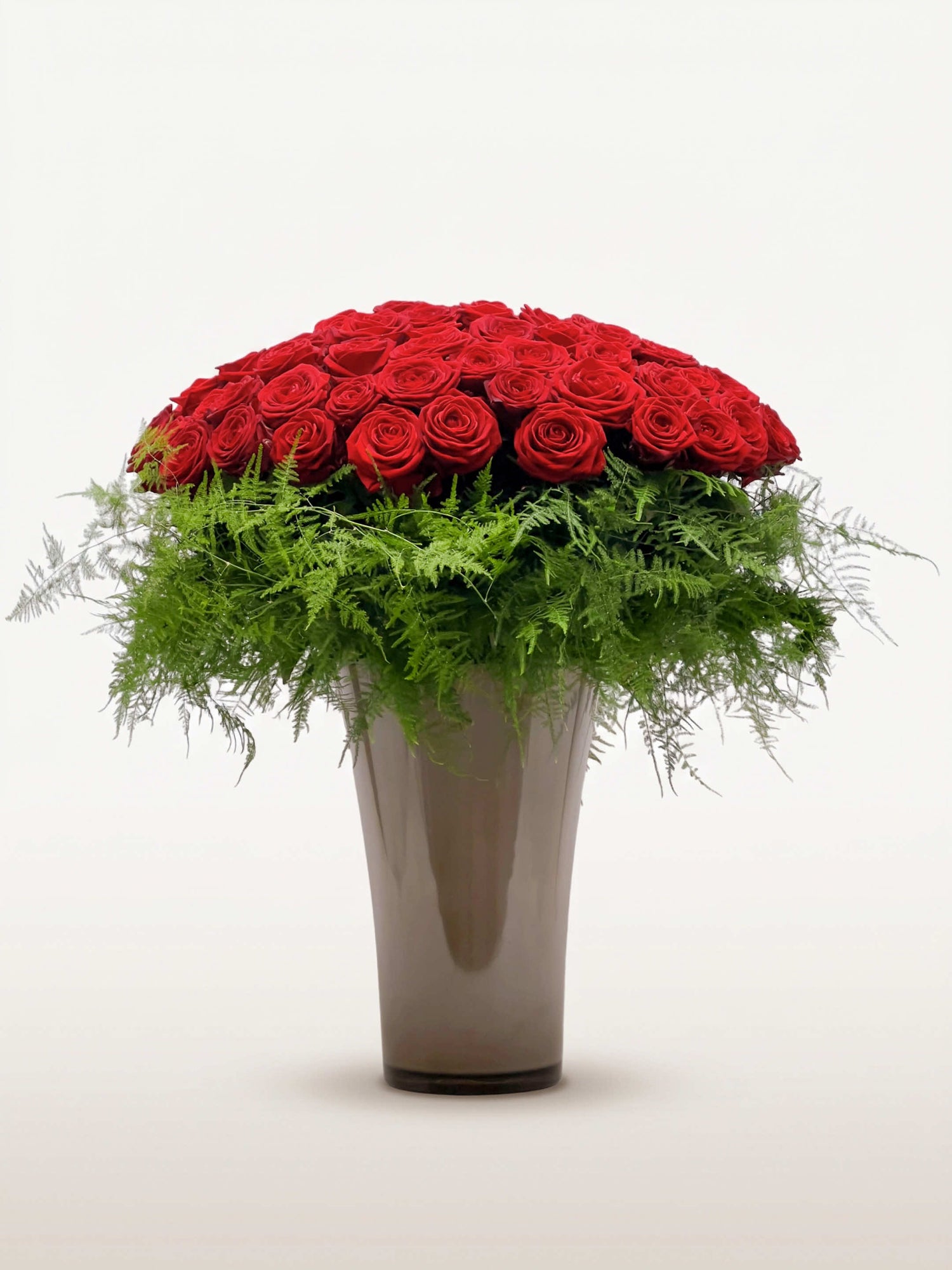 Royal N°100 rassemble une brassée XXL de roses rouges en dôme sur un lit de fougères, dans un vase taupe