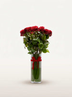 Royal N°25 rassemble une brassée de roses rouges veloutées dans un vase cylindrique en verre, noué d'un ruban rouge