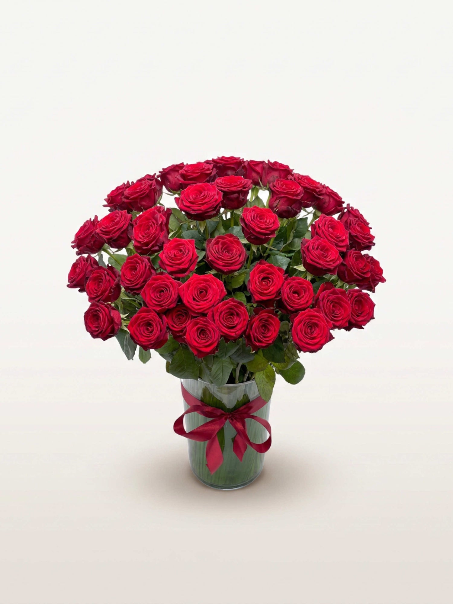 Royal N°75 rassemble une brassée XL de roses rouges veloutées dans un vase en verre noué d'un ruban bordeaux